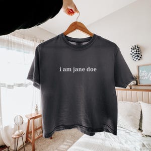 May include: A charcoal grey short-sleeve t-shirt featuring the text i am jane doe printed in white lowercase letters across the centre of the chest. The garment has a crew neckline and a relaxed fit, displayed on a wooden hanger.