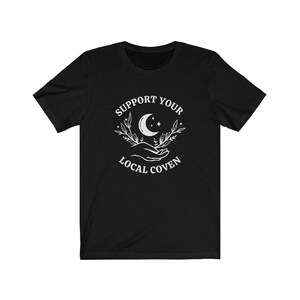 May include: Black t-shirt with a white graphic design that says "Support Your Local Coven" with a crescent moon and stars in the design.