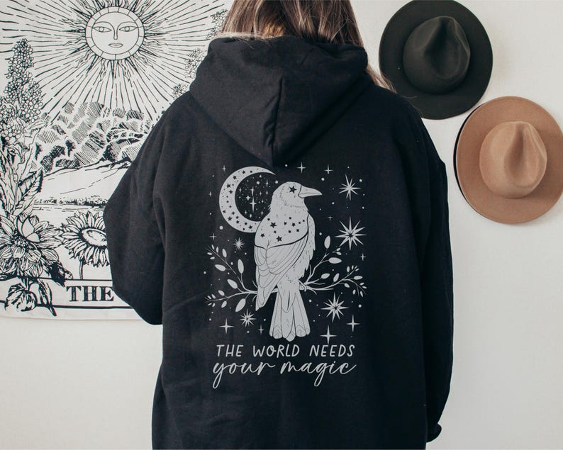 May include: Black hoodie with a graphic of a raven perched on a branch, with a crescent moon and stars. The text "THE WORLD NEEDS your magic" is below the raven. The hoodie is a dark color.