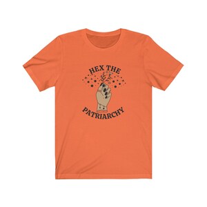May include: Orange t-shirt with the text "HEX THE PATRIARCHY" above an illustration of a hand holding a sprig. The hand has black nail polish and a ring. The design includes stars and is in a vintage style.