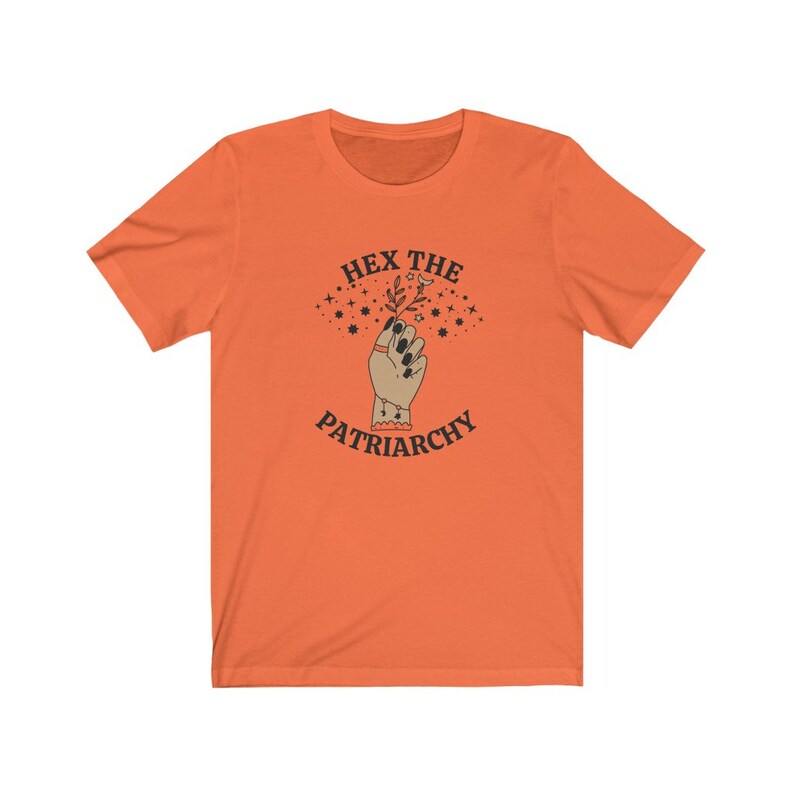 May include: Orange t-shirt with the text "HEX THE PATRIARCHY" above an illustration of a hand holding a sprig. The hand has black nail polish and a ring. The design includes stars and is in a vintage style.