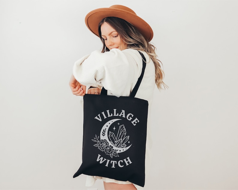 Pode incluir: Bolsa tote preta com a frase "VILLAGE WITCH" em branco. O design inclui uma lua crescente, cristais e elementos florais. A bolsa tem al&ccedil;as pretas e &eacute; segurada por uma pessoa com um chap&eacute;u marrom.