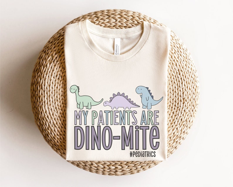 Może przedstawiać: Beżowa koszulka z grafiką przedstawiającą trzy kresk&oacute;wkowe dinozaury i tekstem "Moi pacjenci są dino-mite #Pediatrics".