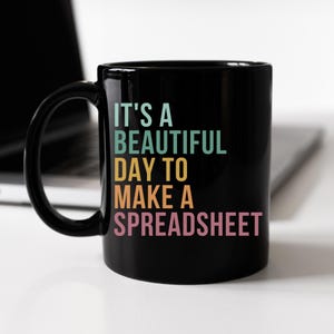 May include: Black ceramic mug with the text "IT'S A BEAUTIFUL DAY TO MAKE A SPREADSHEET" in teal, yellow, and pink. The mug has a large handle and is set against a blurred background.