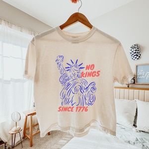 May include: Cream-colored t-shirt with a graphic of the Statue of Liberty in blue and the words "NO KINGS" in pink and "SINCE 1776" in pink. The shirt is hanging on a wooden hanger.
