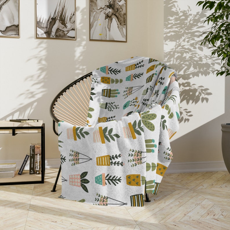 May include: A white throw blanket featuring a repeating pattern of potted plants in various colours, including green, yellow, pink, and blue. The blanket is draped over a chair with a woven rattan frame.