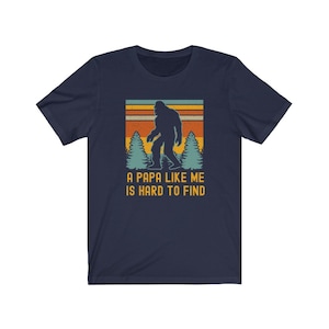 May include: A navy blue t-shirt with a retro-style graphic of Bigfoot walking through a forest. The text "A Papa Like Me Is Hard To Find" is printed below the graphic.