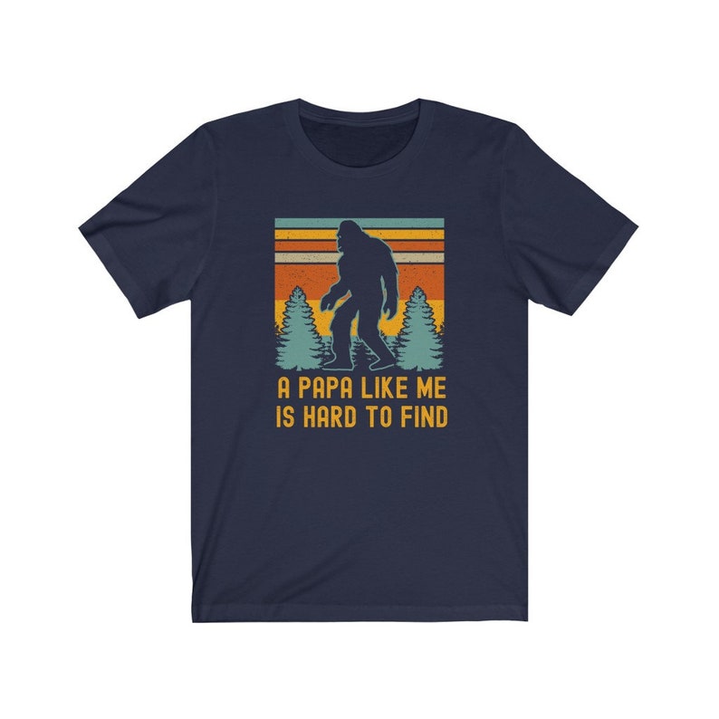 May include: A navy blue t-shirt with a retro-style graphic of Bigfoot walking through a forest. The text "A Papa Like Me Is Hard To Find" is printed below the graphic.