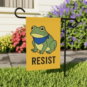 May include: A yellow garden flag features a cartoon frog wearing a blue bandana. The frog is green and yellow, with large eyes. The word "RESIST" is printed in black block letters below the frog. The flag is displayed on a black metal stand in a garden.