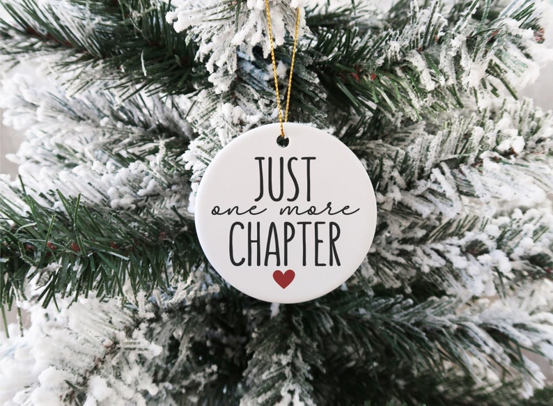 May include: White ceramic Christmas ornament with black text that reads "Just one more CHAPTER" with a small red heart below the text. The ornament is hanging on a snowy artificial Christmas tree.