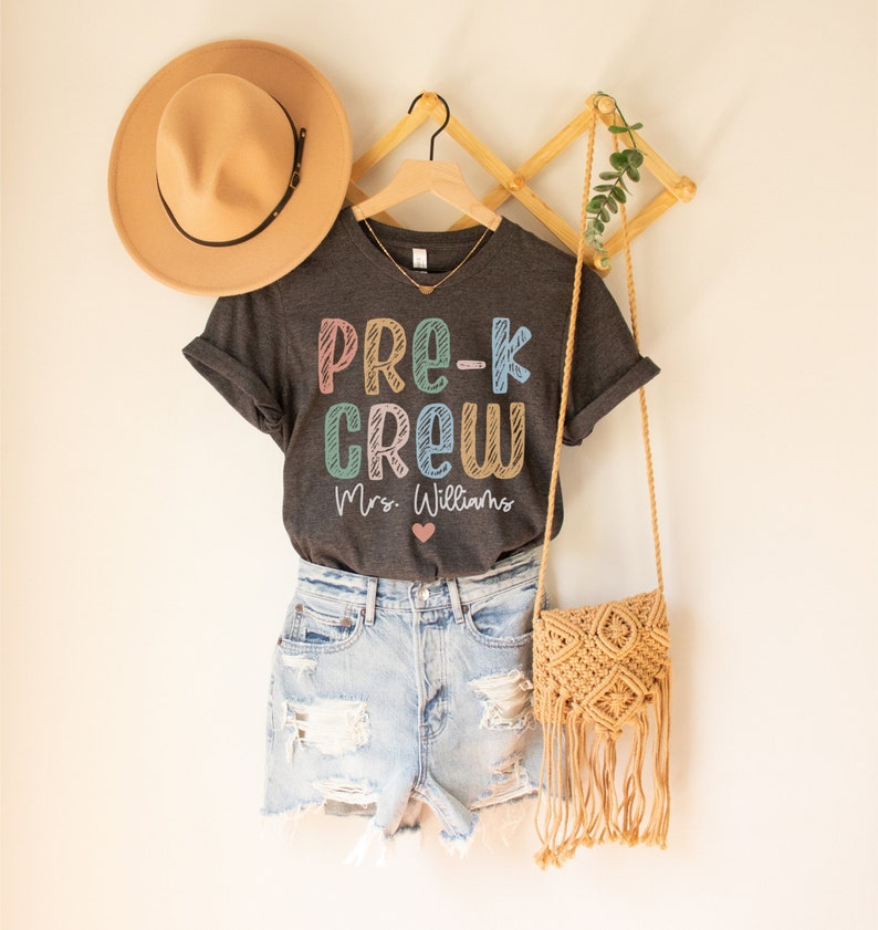 May include: A gray t-shirt with the text "Pre-K Crew" in colorful letters and "Mrs. Williams" below. The shirt is hanging on a wooden hanger with a brown hat and a straw bag.
