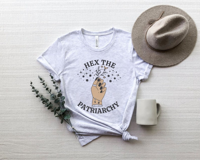 May include: Light gray t-shirt with the text "HEX THE PATRIARCHY" and a graphic of a hand holding a sprig. The design includes stars and a crescent moon. A beige hat and a white mug are next to the shirt.