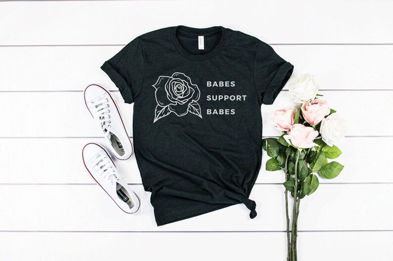 May include: A dark gray t-shirt with a white outline of a rose and the text "BABES SUPPORT BABES".