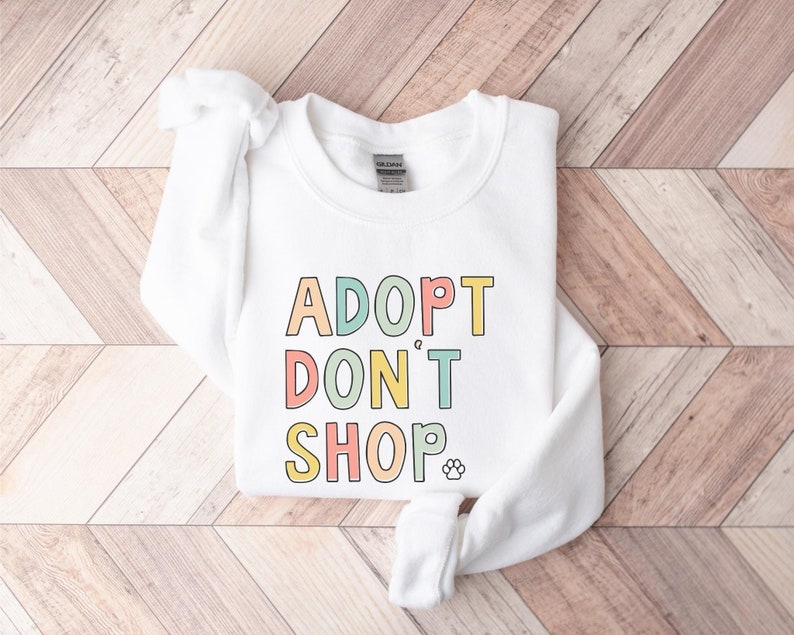 May include: White sweatshirt with the text "ADOPT DON'T SHOP" in rainbow colors with a paw print at the end of the text.