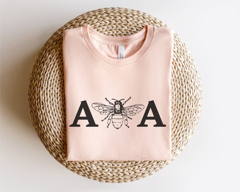 K&ouml;nnte beinhalten: Ein pfirsichfarbenes T-Shirt mit einer schwarzen Biene und den Buchstaben "A & A" auf der Vorderseite.