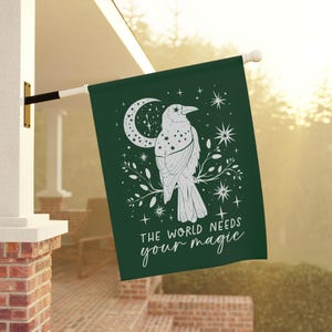 May include: A green garden flag with a white raven silhouette perched on a branch. The raven is surrounded by stars and a crescent moon. The text "The world needs your magic" is printed below the raven.
