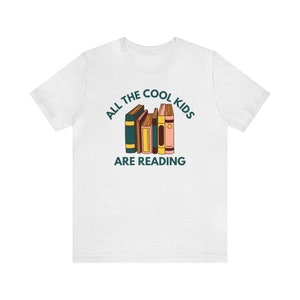 Puede incluir: Camiseta blanca con el texto "All the cool kids are reading" y una pila de libros coloridos.