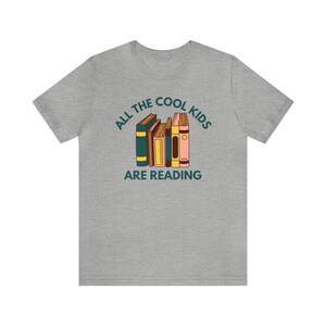 Puede incluir: Camiseta gris con el texto "All the cool kids are reading" y una pila de libros coloridos.