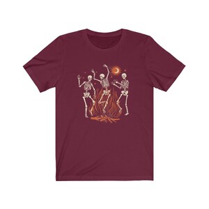 May include: A maroon t-shirt with a graphic of three skeletons dancing around a fire. The skeletons are white with black outlines and the fire is orange and yellow. There is a crescent moon and stars in the background.