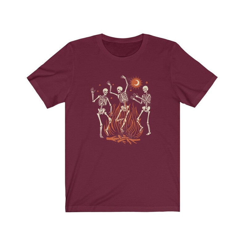 May include: A maroon t-shirt with a graphic of three skeletons dancing around a fire. The skeletons are white with black outlines and the fire is orange and yellow. There is a crescent moon and stars in the background.