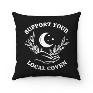 May include: Black throw pillow with white text that reads "Support Your Local Coven". The text is surrounded by a crescent moon, stars, and floral designs.