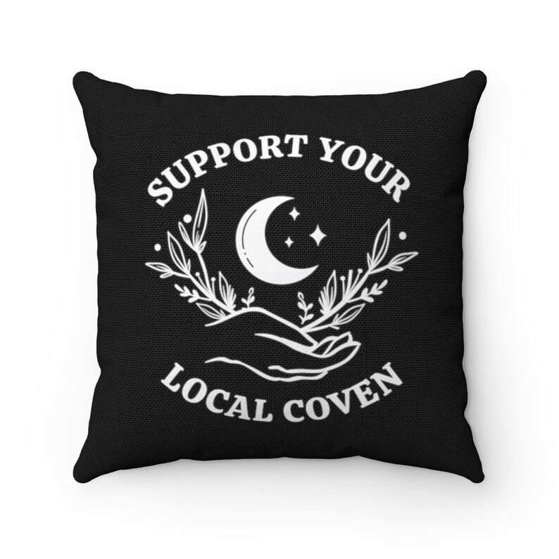 May include: Black throw pillow with white text that reads "Support Your Local Coven". The text is surrounded by a crescent moon, stars, and floral designs.