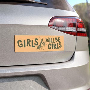 May include: A rectangular bumper sticker with the text "GIRLS WILL BE GIRLS" in dark green letters. A sketch of two praying mantises is between the words. The sticker has a light orange background and is affixed to the back of a silver car.