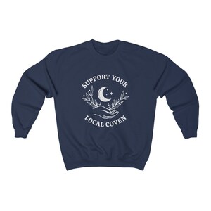 May include: Navy blue sweatshirt with a white graphic of a crescent moon, stars, and two hands holding a sprig of leaves. The text "SUPPORT YOUR LOCAL COVEN" is printed around the graphic.