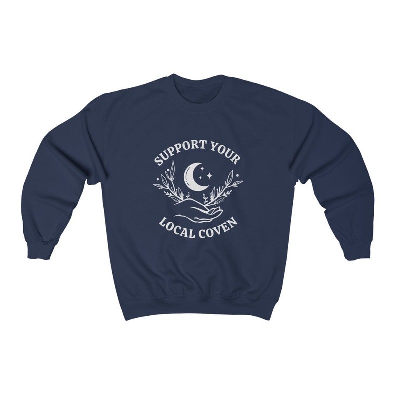 May include: Navy blue sweatshirt with a white graphic of a crescent moon, stars, and two hands holding a sprig of leaves. The text "SUPPORT YOUR LOCAL COVEN" is printed around the graphic.