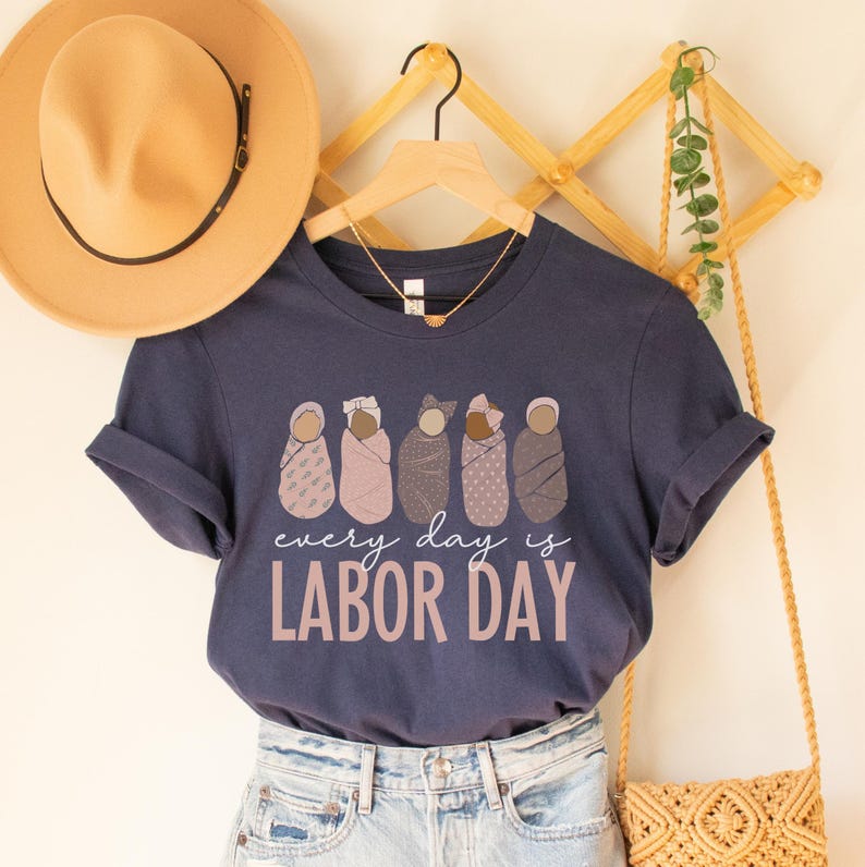 May include: Navy blue t-shirt with the text "every day is LABOR DAY" in a light brown color. The shirt features a graphic of six swaddled babies in various shades of brown and pink. A tan hat and a gold necklace are visible.