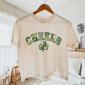 Peut inclure: T-shirt crème avec le mot "CHEERS" en lettres vertes à motifs et un trèfle vert. Le t-shirt est suspendu à un cintre en bois.