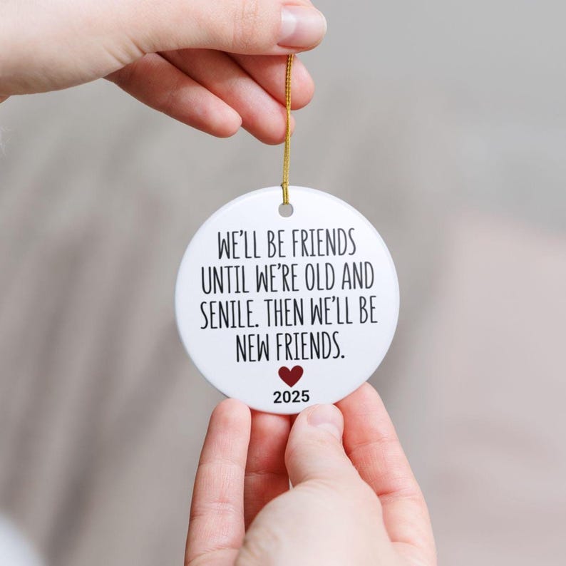 May include: A white, round ornament with a gold string. The ornament has the text "WE'LL BE FRIENDS UNTIL WE'RE OLD AND SENILE. THEN WE'LL BE NEW FRIENDS." with a red heart and the year 2025.