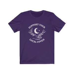 May include: Purple short-sleeved t-shirt with a white graphic design that says "Support Your Local Coven" with a crescent moon and stars in the design.