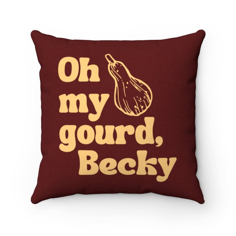 May include: A square throw cushion in a deep maroon colour featuring a light yellow graphic of a butternut squash and the text "Oh my gourd, Becky" printed in a bold, rounded font. The fabric has a visible woven texture throughout the surface.