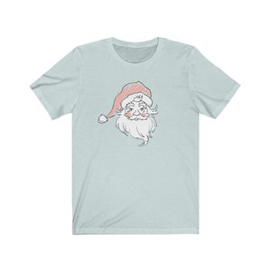 May include: A light blue t-shirt with a vintage-style illustration of Santa Claus's face. The illustration is in shades of pink, white, and gray.