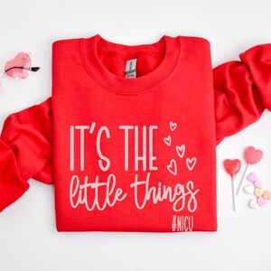 Może przedstawiać: Czerwona bluza z białym napisem "IT'S THE little things", grafiką serca i hashtagiem #NICU. R&oacute;żowe okulary przeciwsłoneczne w kształcie serca i lizaki w kształcie serca r&oacute;wnież znajdują się na zdjęciu.