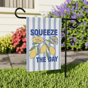 May include: A garden flag with a blue and white striped background. The flag features the words "Squeeze the Day" in blue, along with a lemon illustration. The flag is displayed on a black metal stand in a grassy yard.