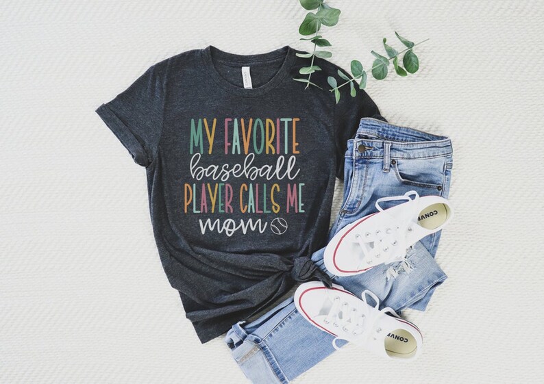 Op de afbeelding: Donkergrijs T-shirt met de tekst "MY FAVORITE baseball PLAYER CALLS ME mom" in kleurrijke letters. Het shirt wordt gecombineerd met een blauwe jeans en witte sneakers, met een honkbalafbeelding. Een casual outfit.