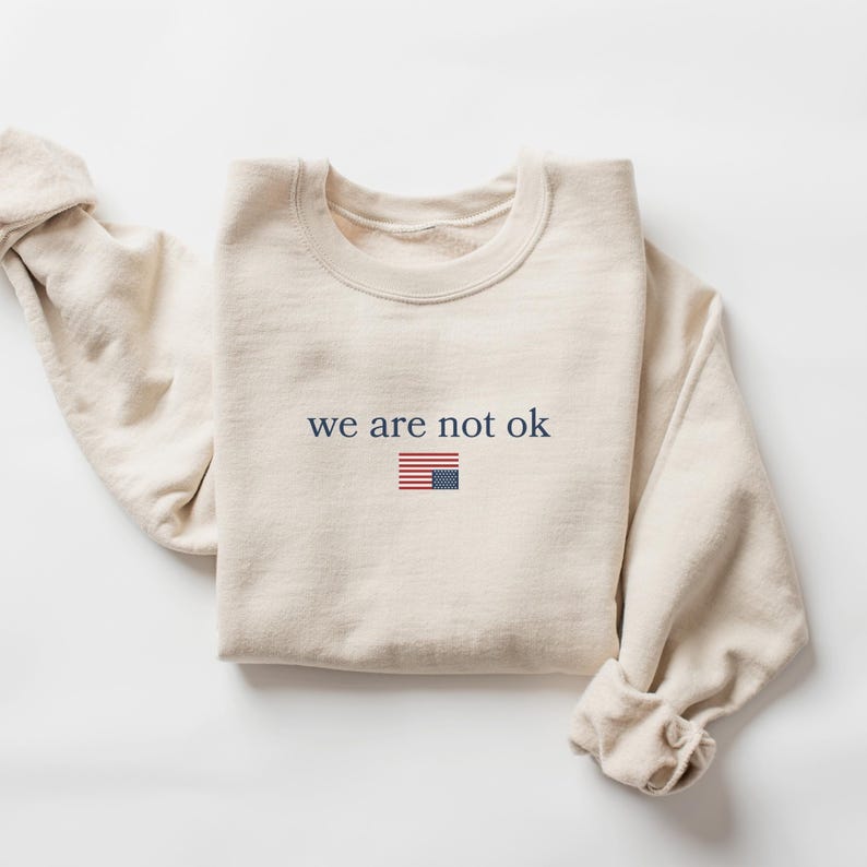 May include: A cream-colored sweatshirt with the text "we are not ok" in navy blue, accompanied by a small American flag graphic. The sweatshirt is folded, with the sleeves casually draped. The overall aesthetic is simple and casual.