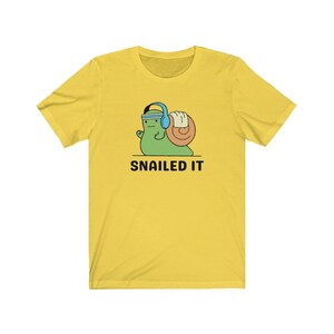 K&ouml;nnte beinhalten: Gelbes T-Shirt mit kurzen &Auml;rmeln, auf dem ein Cartoon-Schnecke mit Kopfh&ouml;rern abgebildet ist, die ein Gewicht hebt. Unter der Schnecke steht der Text "SNAILED IT".