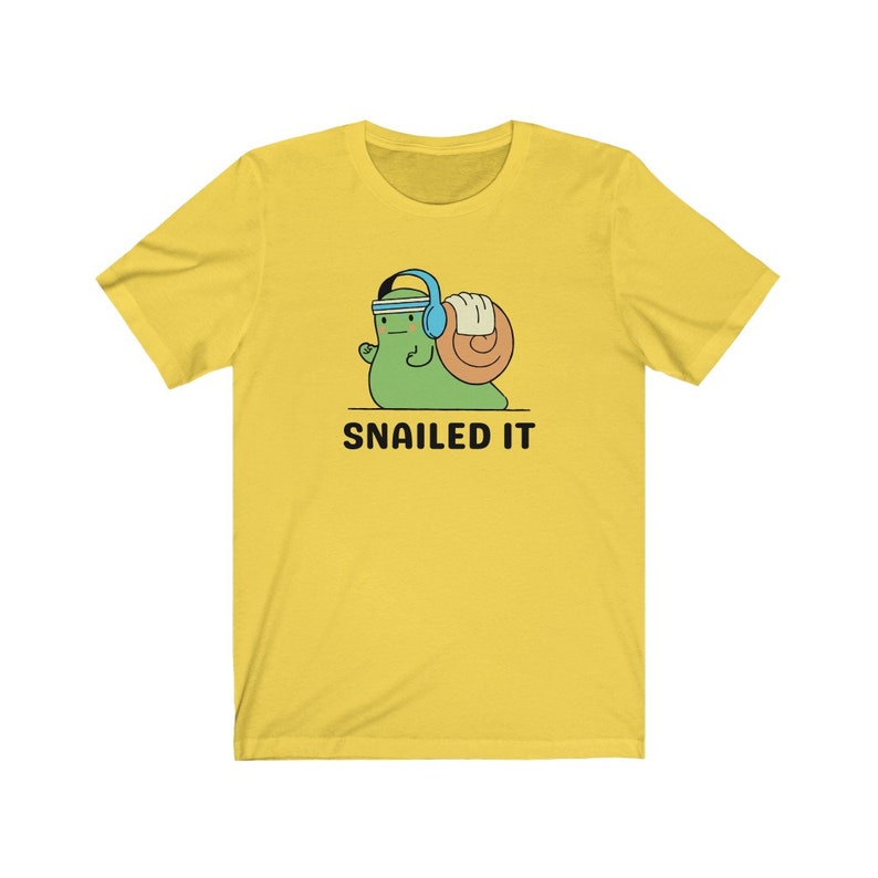 K&ouml;nnte beinhalten: Gelbes T-Shirt mit kurzen &Auml;rmeln, auf dem ein Cartoon-Schnecke mit Kopfh&ouml;rern abgebildet ist, die ein Gewicht hebt. Unter der Schnecke steht der Text "SNAILED IT".