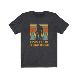 May include: A dark heather grey t-shirt with a vintage-style graphic of a Bigfoot walking through a forest. The text "A Papa Like Me Is Hard To Find" is printed below the graphic.