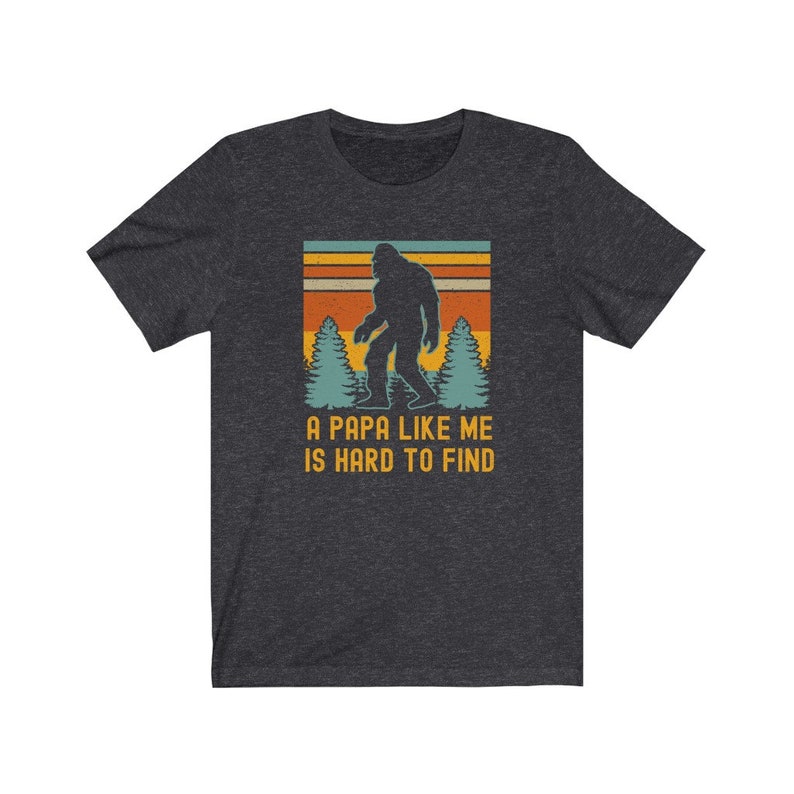 May include: A dark heather grey t-shirt with a vintage-style graphic of a Bigfoot walking through a forest. The text "A Papa Like Me Is Hard To Find" is printed below the graphic.