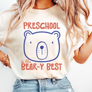 May include: A cream-coloured short-sleeve t-shirt featuring a blue line-art bear face graphic centred on the chest. Above the bear, the text reads PRESCHOOL in red, and below the bear, the text reads is the BEAR-Y BEST in red.