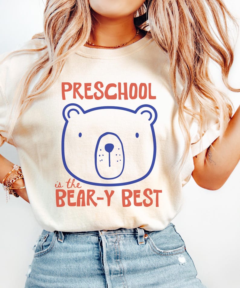 May include: A cream-coloured short-sleeve t-shirt featuring a blue line-art bear face graphic centred on the chest. Above the bear, the text reads PRESCHOOL in red, and below the bear, the text reads is the BEAR-Y BEST in red.