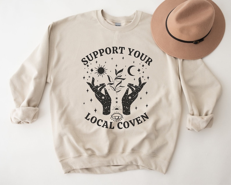 Puede incluir: Una sudadera beige con el texto "Support Your Local Coven" impreso en negro. El dise&ntilde;o incluye un par de manos sosteniendo una planta con una luna, un sol y estrellas.