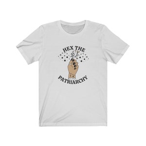 May include: White t-shirt with a graphic design featuring a raised hand holding a sprig of leaves and stars. The text "HEX THE PATRIARCHY" is arched above and below the hand in a circular design.