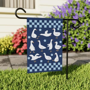 Peut inclure: Un drapeau de jardin bleu avec une bordure à carreaux blancs et un motif d'oies blanches sur fond bleu. Les oies sont dans diverses poses, y compris debout, marchant et volant. Il y a aussi de petites fleurs bleues dispersées dans tout le motif.
