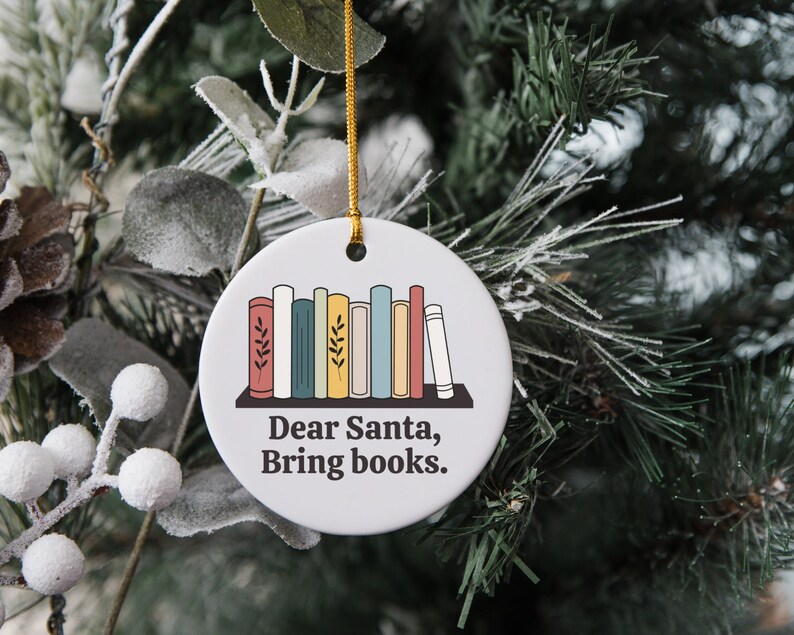 May include: Round white ceramic Christmas ornament with a gold string. The ornament features a bookshelf with colorful books and the text "Dear Santa, Bring books."