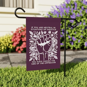 May include: A purple garden flag with white text and a white illustration of a fist surrounded by flowers. The text reads "If you are neutral in situations of injustice you have chosen the side of the oppressor."